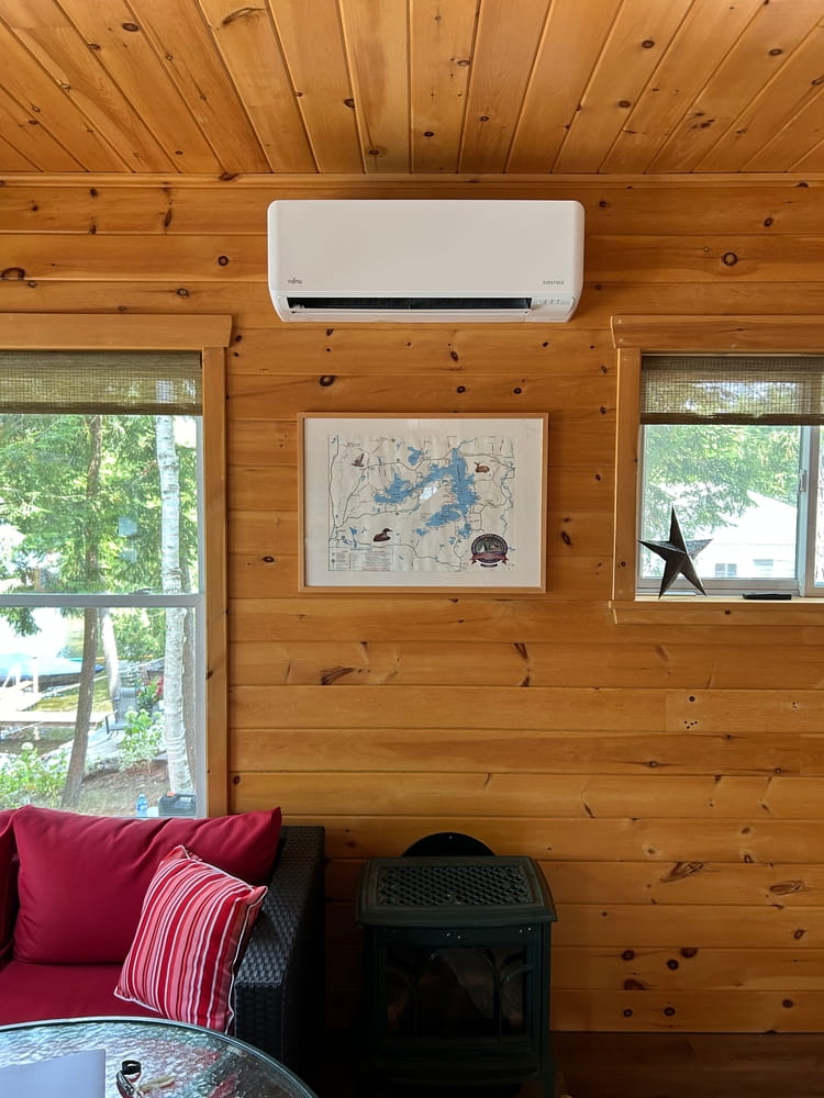 A ductless mini-split installed in a wooden cabin, with map on the wall, sofa and windows visible.